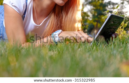 Student is using digital tablet PC in the park lying on the grass. Mobile style of life. Mobile communications. Remote learning or education concept.  Springtime relax. Girl is texting on smart phone.