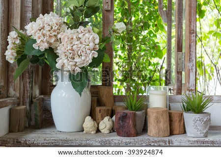 White flower vase and small succulent plants with decoration items placed on a wooden shelf near the windows in rustic atmosphere.