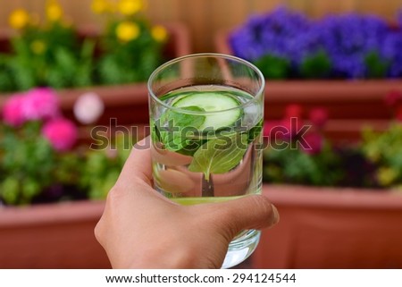 A hand holding a glass of cucumber lime mint Infused water with blurred flowers background