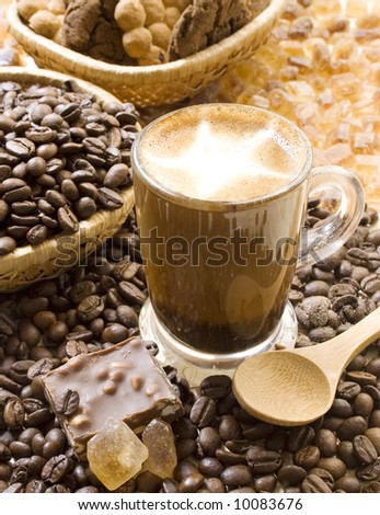 Cup of cappuccino coffee with cinnamon powder, surrounded by coffee beans, cookies, crystallized brown sugar and chocolate.