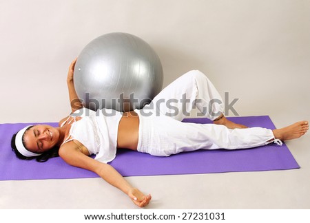 A young woman dressed on white resting after a gym session with a gray gym ball on a lilac mat.