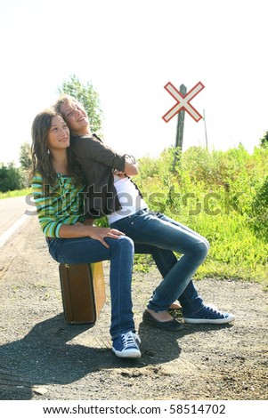 Two teenager girls sit on a old luggage on a road with a rail sign in background