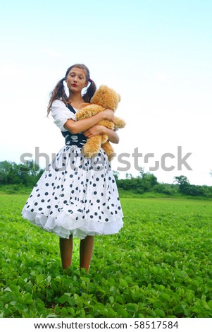 Young girl dress like a doll with polka dots dress having a generic teddy bear in hand and standing in a middle of a green meadow