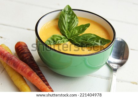 Carrot soup with basil leave in a green bowl with spoon and fresh carrots on a white wooden background