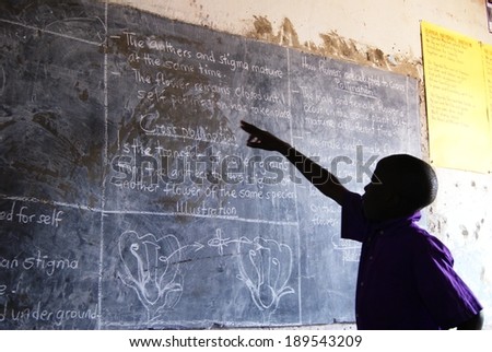 MATUGGA, UGANDA, AFRICA - CIRCA AUGUST 2013: Unidentified young boy in a purple school uniform pointing at the blackboard, reading out loud what he learned today. North of Kampala, Uganda, East Africa