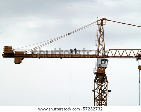 Workers atop a multistory construction crane.  Each is engaged in a different form of communication ... one on a cell phone, the other s hauling something up by rope