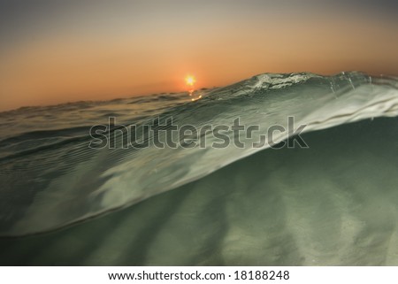 A wave passes by the camera half in, half out of the water while the sun sets on the horizon and the ripples of the ocean floor reflect off the surface.