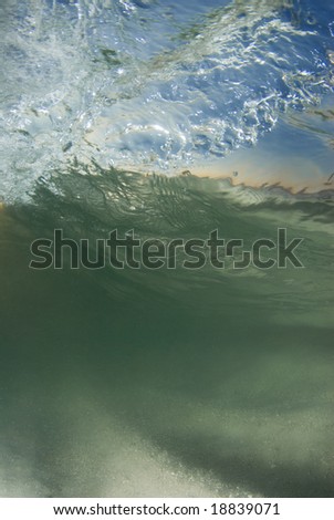 Underwater view of a curling, breaking wave; With the bright blue sky and sunset on the horizon coming through the surface and the stirring sand on the ocean floor
