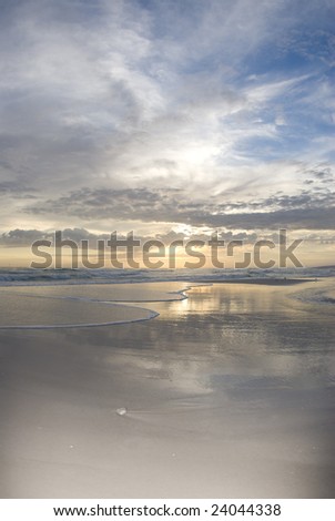 The setting sun reflects off the water as the surf flows in and the clouds glow on the horizon