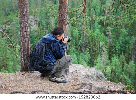 The photographer is sitting on the top of hill against the taiga forest. The tourist is taking pictures.