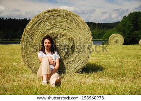 Happy woman is sitting down under the big straw bale. She is resting against the country landscape with field gathered.