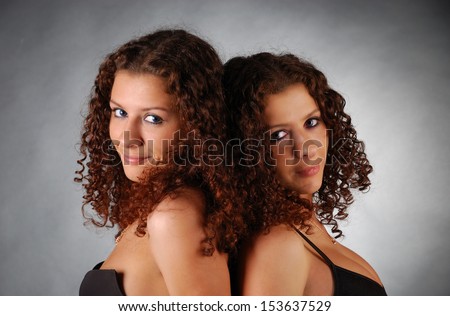 Two young women are clinging close to one another back to back and looking at the camera. Pretty girls are very much alike because they are twins.