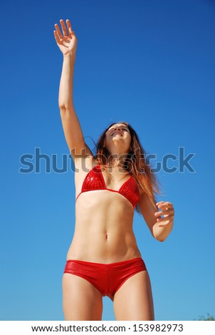 Young woman is waving hand. She is wearing red bikini. Her slender body is photographed from below. She is playing against the blue summer sky. She is acquiring a tan in the sun.