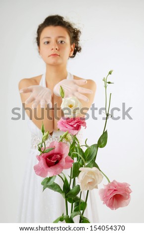 Pretty woman is standing and reaching out both hands in gloves. In the foreground there are gentle flowers. White low-necked dress accentuates her beautiful shoulders and hands.