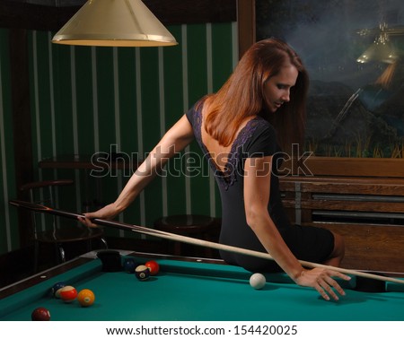 Woman is sitting on the edge of a billiard-table. She is aiming a cue from her back.