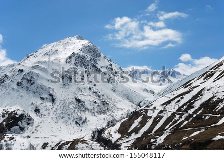 Black-and-white slope with serpentine road is against the mountains snow-covered and azure cloudy sky (the pass of Saint Bernard)
