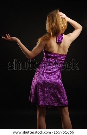 Teenage blonde girl is standing back in the dark. She is wearing a velvet cocktail dress.