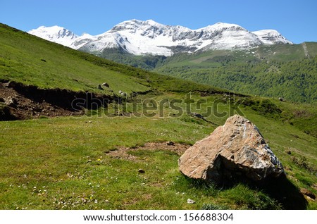 A country road is twisting on the green pasture between the glacis forested and steep slope covered with snow in the spring mountains Pyrenees.