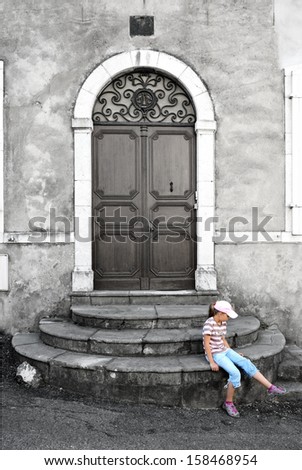 A modern preteen girl is waiting under the closed door. She is sitting on the stone stairs of the old house.
