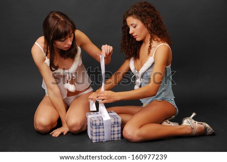 Pretty women are sitting and preparing a christmas gift on the dark background. They are wearing transparent tops and shorts decorated with white fluff.