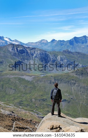 The tourist is standing back on the top of mountain against the green slopes in summer Pyrenees. There are remote peaks with glaciers and the blue sky in the background.
