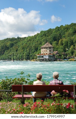A senior couple is sitting on the bench and seeing the sights of Rhine Falls. There are also viewing platforms built on both sides of the Rhine.