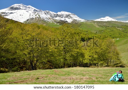 A preteen girl is resting in the green slope. There are spring forest and snow remote mountains.