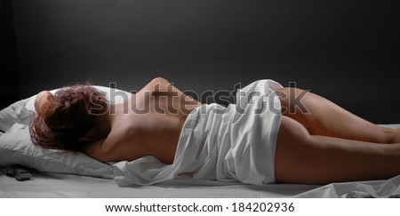 A nude young woman is lying on the white bed-clothes. Her handgun is hidden under the pillow.