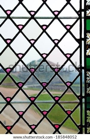 Focus is on the surface of the vintage window of the ancient French castle. There are blurred buildings, lawns and paths through the stained glass.