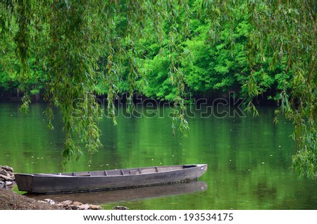 Wood boat is landing near the shore of the pond. Lush foliage are reflected in the calm water surface.