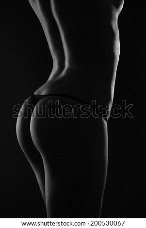 Naked woman is standing back. Her sexy body is photographed in the dark with few highlights. She is wearing tiny black panties.