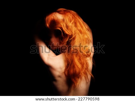 A ginger nude woman is standing back in the dark. She has raised her long red hair and turned left her face.