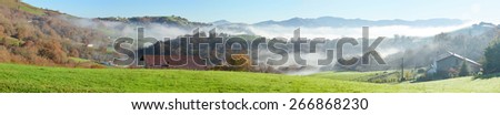 The farms with a green pasture are situated on the forested mountain in the Pays Basque. The slopes are shrouded with mist in the winter sunny day.