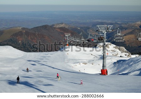 People are being transported with a chair lift up the snowy slope. Another skiers are sliding down on the piste in the winter mountains.
