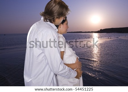 Lovely young couple expecting  a baby standing in profile,embraced and looking together at sunrise and how start a new day at sea on beach