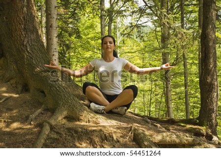 Young woman doing yoga in nature and sitting in lotus position near a tree in forest,breathing the clean air of mountains