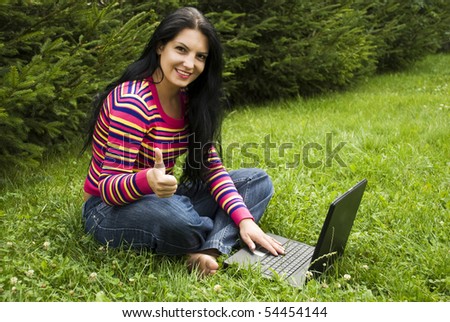 Woman working on laptop and enjoying her vacation in nature at mountain in the middle of fir tress and sitting with legs crossed in green grass and giving thumbs up