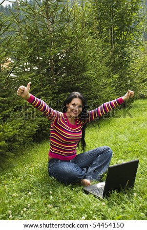 Beautiful happy woman sitting with legs crossed  and working on laptop in vacation at mountain with fir trees around her,she had a successful  business and open hands and giving thumbs up