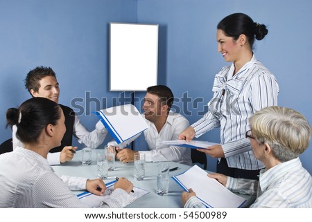 Group of people at meeting having fun and laughing while a business woman giving all folders with papers,blank chart for presentation in background