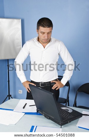Worried business man in office standing with with arms akimbo and looking to laptop