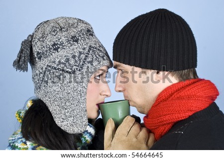 Couple of young people dressed in winter clothes with hats on heads,sweaters standing face to face and looked at each other drinking a hot drink from same mug