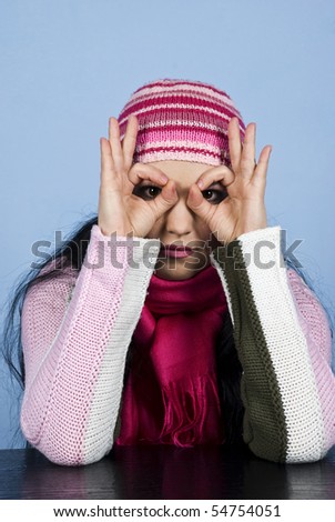 Young woman with cap and pullover standing at table with hands over eyes and looking you through fingers