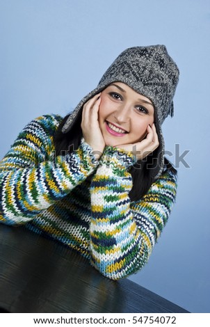Girl in winter cap and pullover sitting  at table smiling and holding hands on face,right angle view over blue background