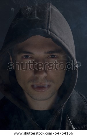 Young man  dressed in black clothes with hood in darkness taking smoke out on mouth