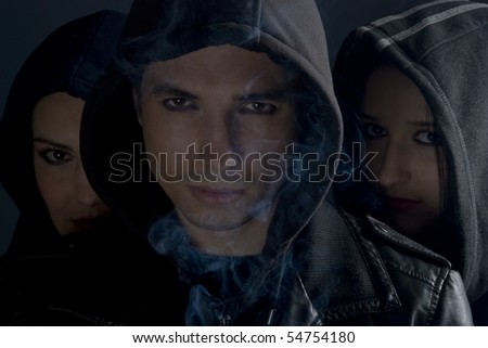 Urban people street in black hoods standing in darkness  with smoke around them and looking you.man in front and two women behind him