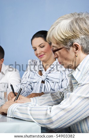 Senior businesswoman with eyeglasses write on paperwork at meeting and her young colleague smile and writing also,focus on older woman