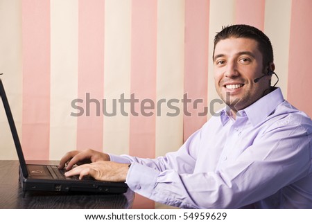 Call center man working on laptop in office and smiling at you,vertical blinds background