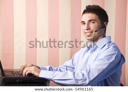 Happy helpdesk young male sitting at desktop and typing at laptop looking you and smile,copy space for text message in left part of image