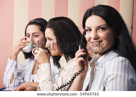 Three business women at office,first woman speaking at phone and smiling and the other drinking coffee and having an conversation