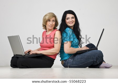 Two women sitting back to back on parquet and both using laptop,every woman looks over shoulder at the other and having an coversation and smiling